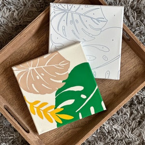May include: Two square canvases with botanical designs rest on a wooden tray. One canvas features a green and tan leaf design, while the other has a line drawing of a leaf. The tray is light brown.
