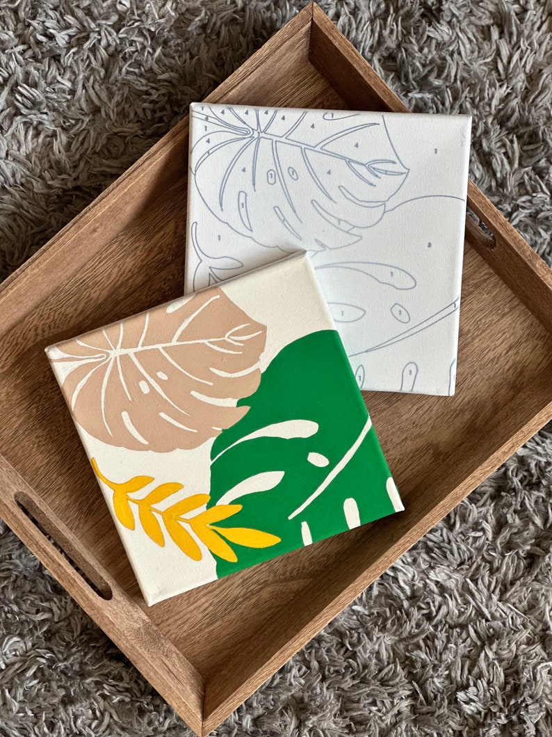 May include: Two square canvases with botanical designs rest on a wooden tray. One canvas features a green and tan leaf design, while the other has a line drawing of a leaf. The tray is light brown.