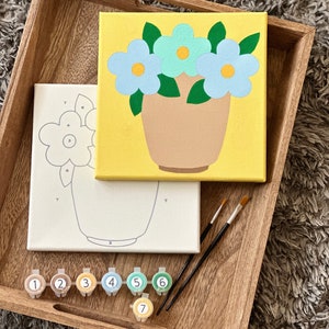 May include: A paint by numbers kit with a canvas featuring a yellow background with a brown pot and three blue flowers. The kit includes seven paint pots with numbers 1 through 7 and two paintbrushes.