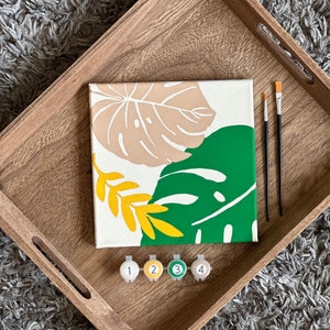 May include: A paint-by-numbers kit featuring a canvas with a botanical design in beige, green, and yellow. The kit includes paintbrushes and numbered paint pots, all arranged on a wooden tray.