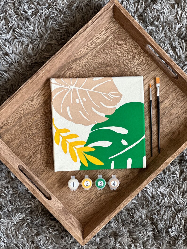 May include: A paint-by-numbers kit featuring a canvas with a botanical design in beige, green, and yellow. The kit includes paintbrushes and numbered paint pots, all arranged on a wooden tray.