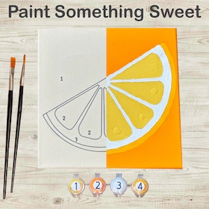 May include: A paint-by-numbers kit featuring a lemon slice design. The canvas is split, with one side showing the numbered outline and the other side painted. The kit includes paintbrushes and numbered paint pots. The text "Paint Something Sweet" is at the top.