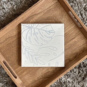 May include: A square paint-by-numbers canvas featuring a tropical leaf design with numbered sections. The canvas is set inside a light brown wooden tray. The background is a gray, textured rug.