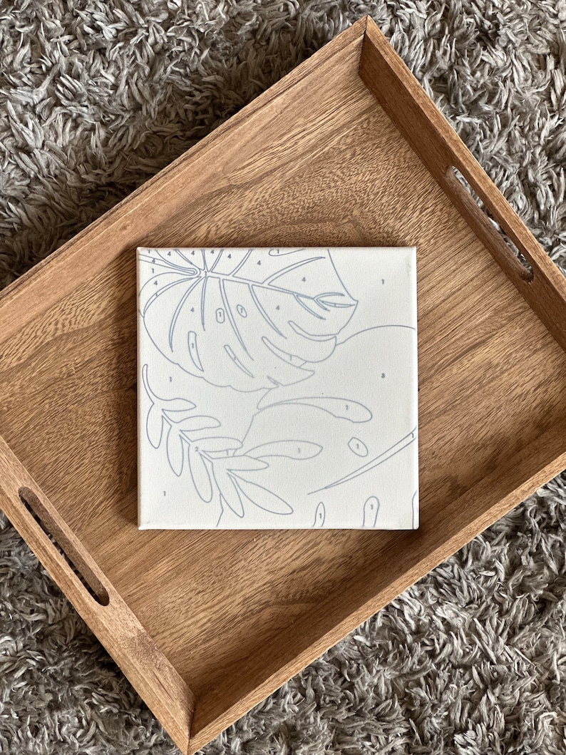 May include: A square paint-by-numbers canvas featuring a tropical leaf design with numbered sections. The canvas is set inside a light brown wooden tray. The background is a gray, textured rug.