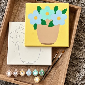 May include: A paint-by-number kit featuring a square canvas with a finished painting of light blue and mint green flowers in a tan vase against a yellow background, an unpainted numbered canvas, two black paintbrushes, and seven numbered paint pots.
