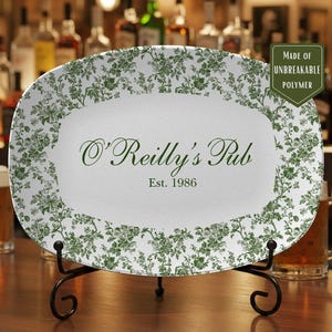 May include: White oval serving platter with green floral design and the text "O'Reilly's Pub Est. 1986". The platter is made of unbreakable polymer and is displayed on a black metal stand. The background shows a bar setting.