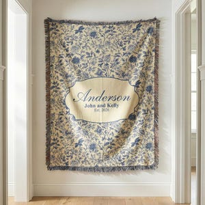 May include: A cream-colored throw blanket with a blue floral pattern and a decorative border. The blanket features a central cream-colored label with the name "Anderson", and the names "John and Kelly" and the year "Est. 2026".