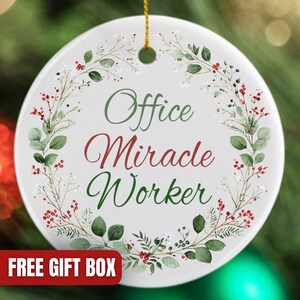 May include: A white ceramic Christmas ornament with the words "Office Miracle Worker" in green and red script, surrounded by a wreath of green leaves and red berries. A gold string is attached for hanging.