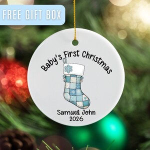May include: A white ceramic Christmas ornament with a blue and white patchwork stocking design. The text "Baby's First Christmas" and the name "Samuel John" with the year "2026" are printed on the ornament.