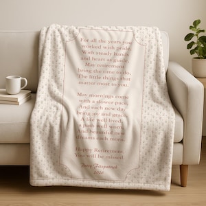 May include: A cream-colored throw blanket with a floral pattern and a framed message in a soft red hue. The text reads "Happy Retirement. You will be missed." and includes a name and year.