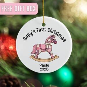 May include: A white ceramic Christmas ornament with a pink and purple rocking horse design. The text "Baby's First Christmas" and the name "Paige" with the year "2026" are printed on the ornament. A gold string is attached for hanging.
