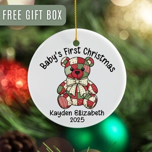 May include: A white ceramic Christmas ornament with a patchwork teddy bear design. The ornament reads "Baby's First Christmas" and includes a name and the year 2025. A gold string is attached for hanging. The words "FREE GIFT BOX" are in the top left corner.