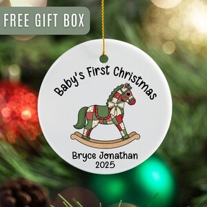 May include: A white ceramic Christmas ornament with a rocking horse design. The ornament reads "Baby's First Christmas" and includes a name and year. The ornament is hanging against a backdrop of Christmas decorations.