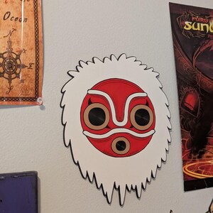 May include: A white and red mask with a black outline, featuring a central red circle and brown eye-like shapes. The mask has a white, furry-like border. The mask is mounted on a light gray wall, with other artwork visible in the background.