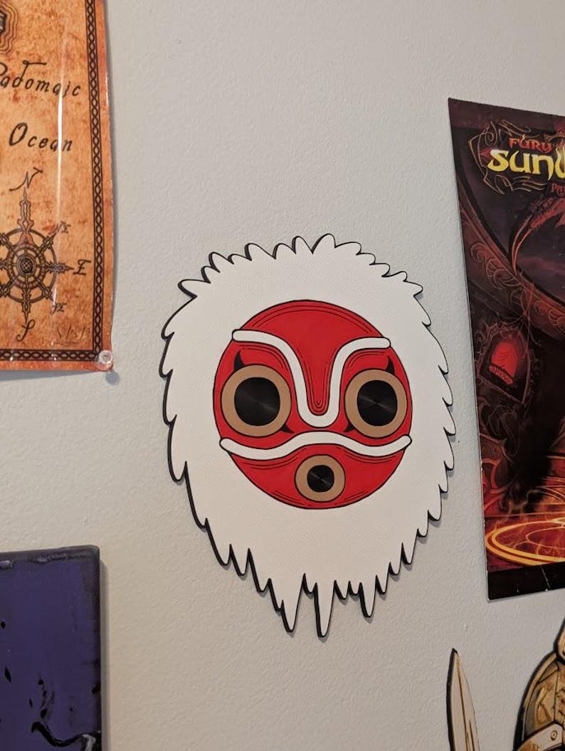 May include: A white and red mask with a black outline, featuring a central red circle and brown eye-like shapes. The mask has a white, furry-like border. The mask is mounted on a light gray wall, with other artwork visible in the background.