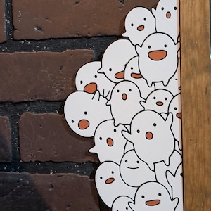 May include: A wooden framed print featuring a collection of white cartoon ghosts with orange mouths and black eyes. The background is a brick wall. The ghosts are clustered together, some peeking out from behind the frame.