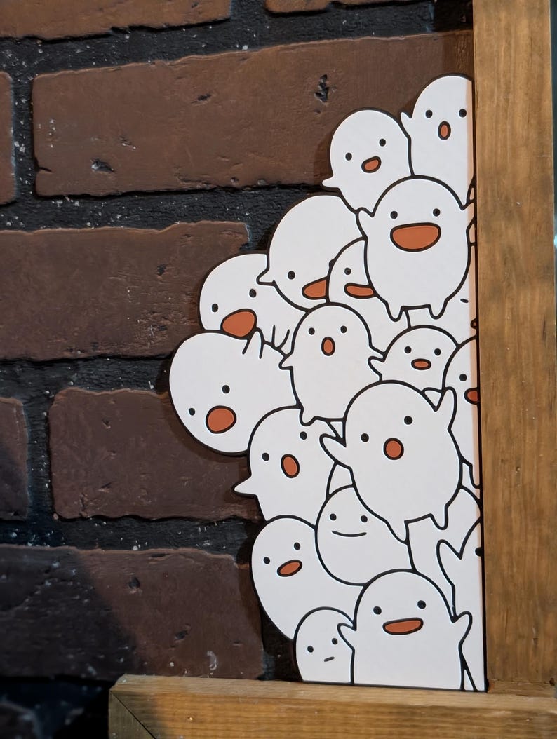 May include: A wooden framed print featuring a collection of white cartoon ghosts with orange mouths and black eyes. The background is a brick wall. The ghosts are clustered together, some peeking out from behind the frame.