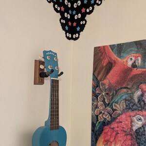 May include: A bright blue Makala ukulele hangs on a wooden wall mount. Above it, a black corner wall decal features a repeating pattern of white cartoon eyes, pink stars, and light blue stars, following the angle of the wall corner.