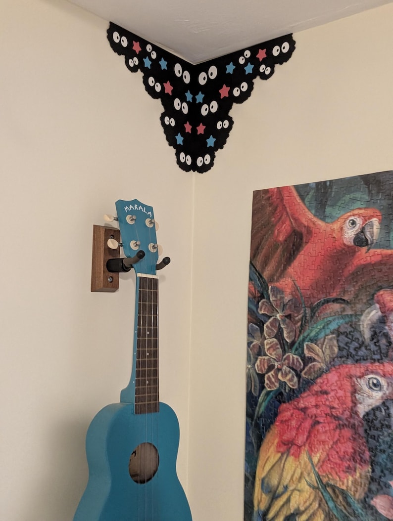 May include: A bright blue Makala ukulele hangs on a wooden wall mount. Above it, a black corner wall decal features a repeating pattern of white cartoon eyes, pink stars, and light blue stars, following the angle of the wall corner.