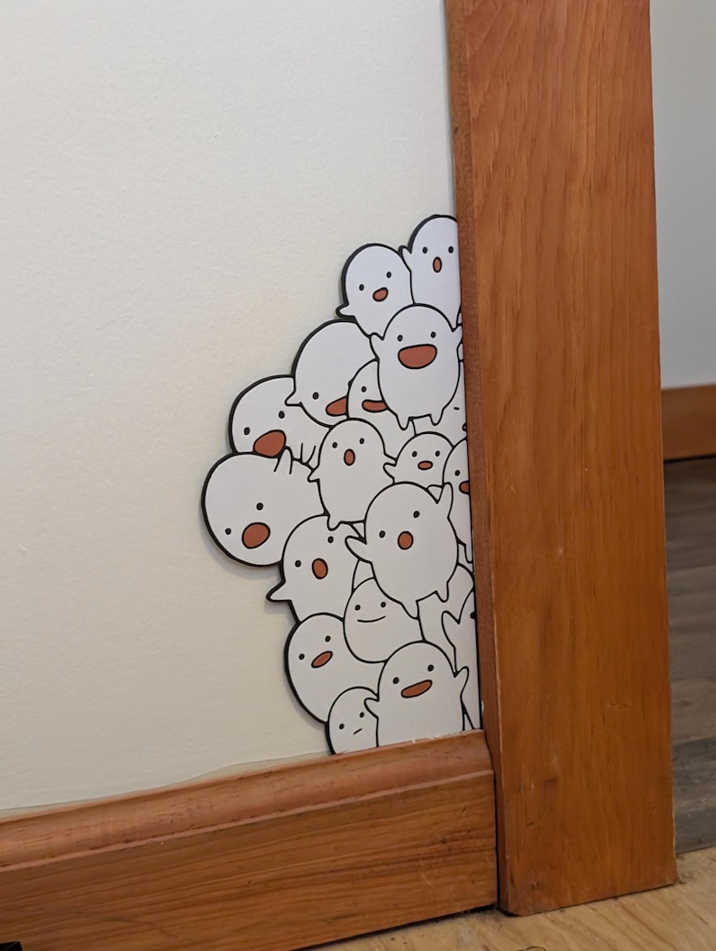 May include: A cluster of white ghost stickers with black outlines and small red accents. The stickers are placed on a white wall, partially obscured by a wooden frame. The ghosts have various expressions.