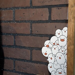 May include: A collection of white cartoon ghost stickers with orange mouths and black eyes. The stickers are clustered together, peeking out from behind a wooden frame. The background features a brick wall with dark brown bricks.