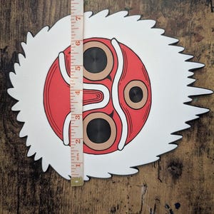 May include: A decorative mask featuring a red and white design with black accents. The mask has a circular face with two brown eye shapes and a white, jagged border. The mask measures approximately 7 inches in height.