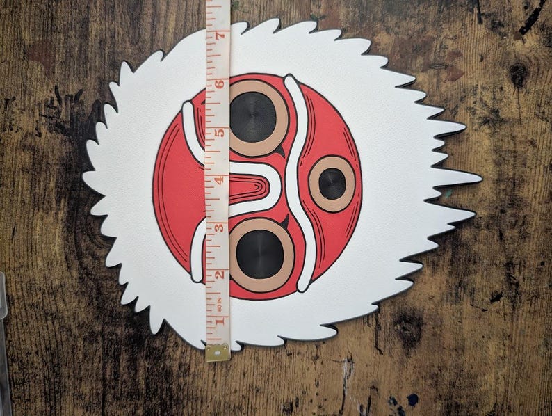 May include: A decorative mask featuring a red and white design with black accents. The mask has a circular face with two brown eye shapes and a white, jagged border. The mask measures approximately 7 inches in height.