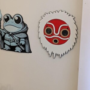 May include: Two cartoon-style magnets. One is a frog wizard in a cloak, holding a staff. The other is a white, red, and brown mask with a jagged edge, reminiscent of a character from a Japanese animated film.