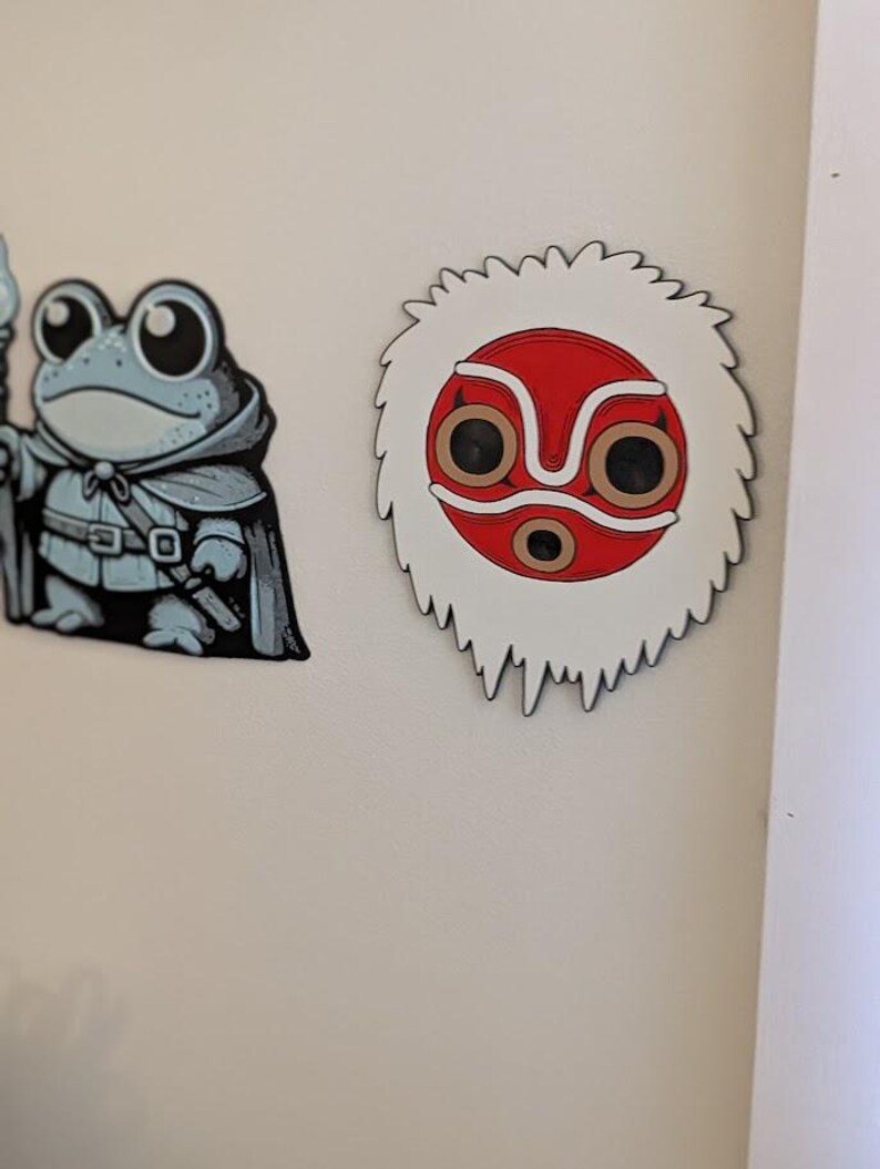 May include: Two cartoon-style magnets. One is a frog wizard in a cloak, holding a staff. The other is a white, red, and brown mask with a jagged edge, reminiscent of a character from a Japanese animated film.