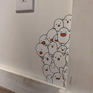 May include: A cluster of white ghost stickers with black eyes and orange mouths are affixed to a white wall. The stickers are positioned around a corner, with some visible on the adjacent wall. A white electrical outlet is visible.
