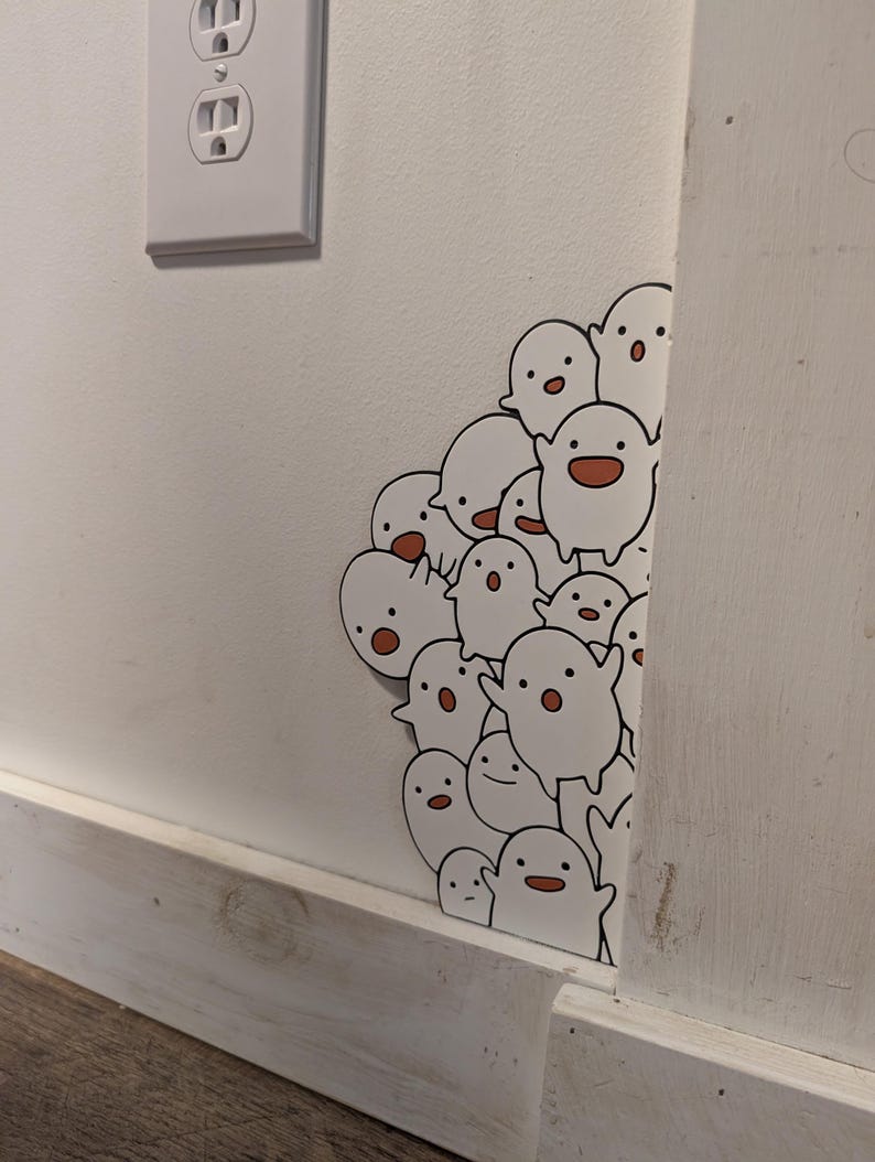 May include: A cluster of white ghost stickers with black eyes and orange mouths are affixed to a white wall. The stickers are positioned around a corner, with some visible on the adjacent wall. A white electrical outlet is visible.