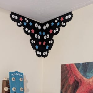 May include: A black corner wall decal featuring a scalloped edge, multiple pairs of white eyes with black pupils, and scattered small red and blue stars. The decal is applied to the upper corner of a room, positioned above a blue Makala ukulele.