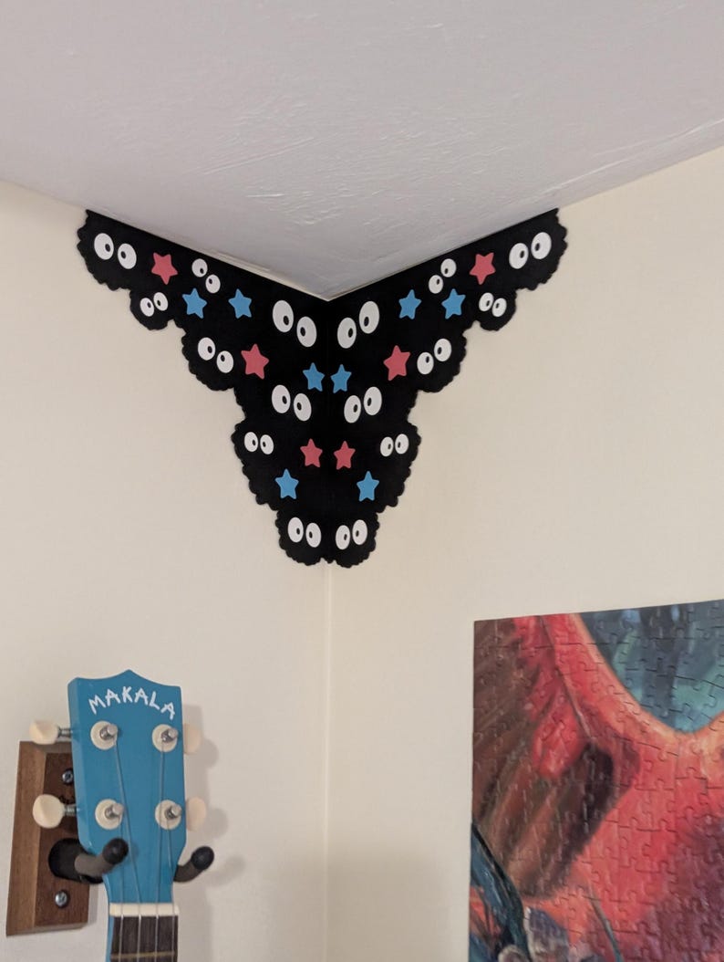 May include: A black corner wall decal featuring a scalloped edge, multiple pairs of white eyes with black pupils, and scattered small red and blue stars. The decal is applied to the upper corner of a room, positioned above a blue Makala ukulele.
