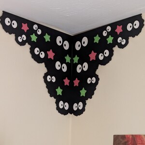 May include: A black corner wall decal featuring a scalloped edge design with multiple pairs of white eyes and scattered small pink and green stars. The decal is applied to the intersection of two white walls, creating a decorative corner accent.