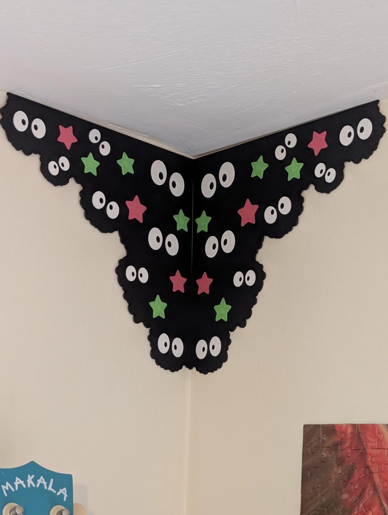 May include: A black corner wall decal featuring a scalloped edge design with multiple pairs of white eyes and scattered small pink and green stars. The decal is applied to the intersection of two white walls, creating a decorative corner accent.