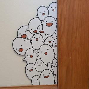 May include: A cluster of white ghost stickers with black outlines and smiling faces with brown mouths. The stickers are on a white wall, partially obscured by a wooden door frame.