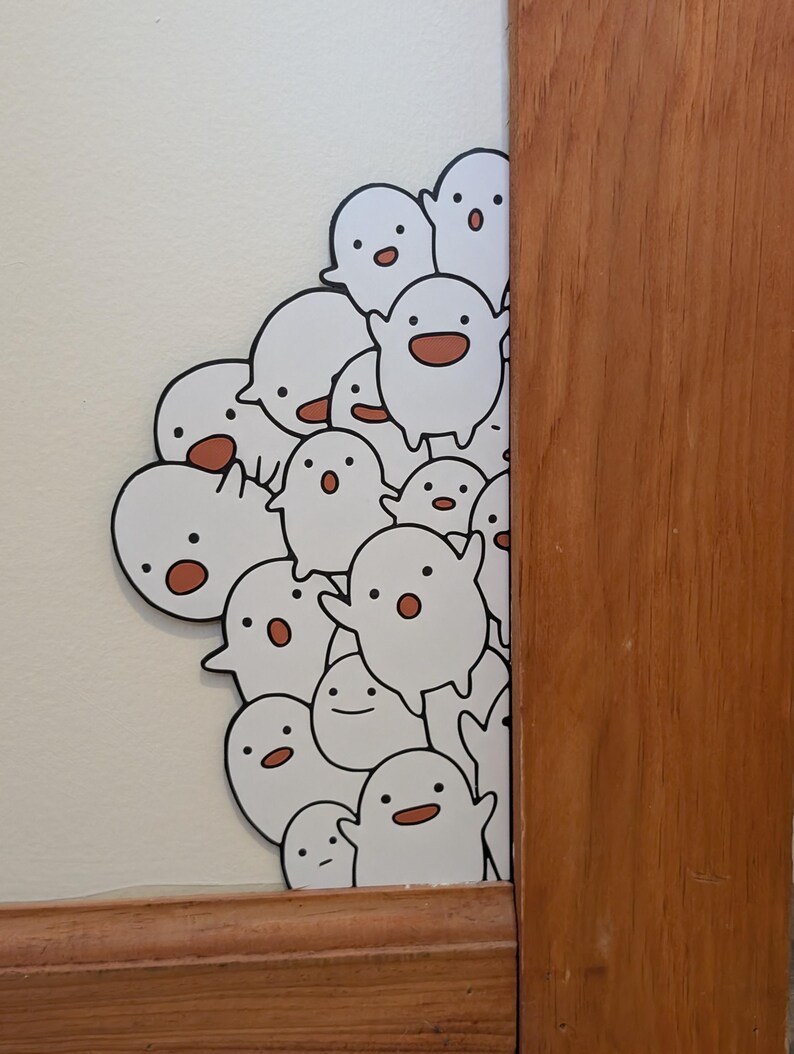 May include: A cluster of white ghost stickers with black outlines and smiling faces with brown mouths. The stickers are on a white wall, partially obscured by a wooden door frame.
