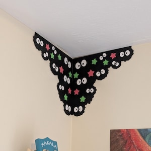 May include: A black corner wall decal featuring multiple pairs of white eyes with black pupils, interspersed with small pink and green star shapes. The decal is shaped to fit into the corner where two walls meet, creating a decorative accent.