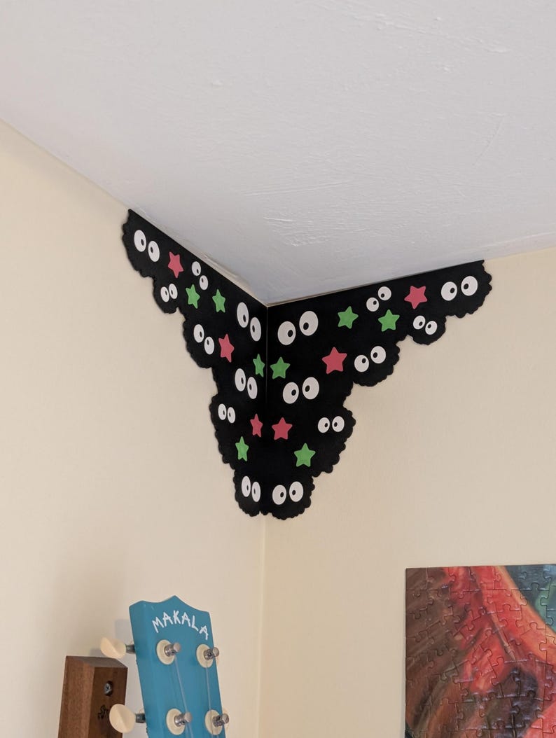 May include: A black corner wall decal featuring multiple pairs of white eyes with black pupils, interspersed with small pink and green star shapes. The decal is shaped to fit into the corner where two walls meet, creating a decorative accent.