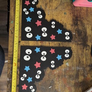 May include: Two black L-shaped foam pieces decorated with white cartoon eyes and red and blue stars. A yellow measuring tape is placed vertically along the left side, showing the pieces are approximately 17 inches in total length.