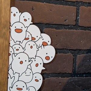 May include: A white artwork featuring a group of cartoon ghosts with orange mouths and eyes. The artwork is set against a brick wall and framed in wood.