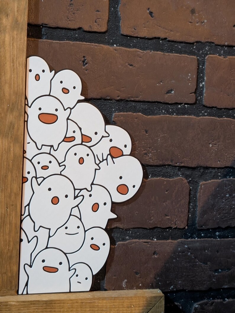 May include: A white artwork featuring a group of cartoon ghosts with orange mouths and eyes. The artwork is set against a brick wall and framed in wood.