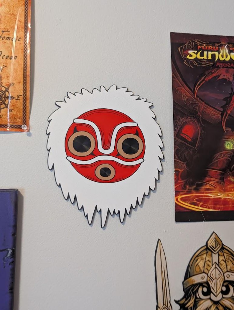 May include: A red and white mask with a stylized design, featuring large circular eye openings and a white, fringed border. The mask is mounted on a light gray wall, with other artwork visible in the background.