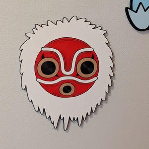 May include: A decorative wooden mask featuring a red face with white and brown accents, and black eye sockets. The mask is surrounded by a white, furry border with a jagged edge. The mask is mounted on a light gray wall.