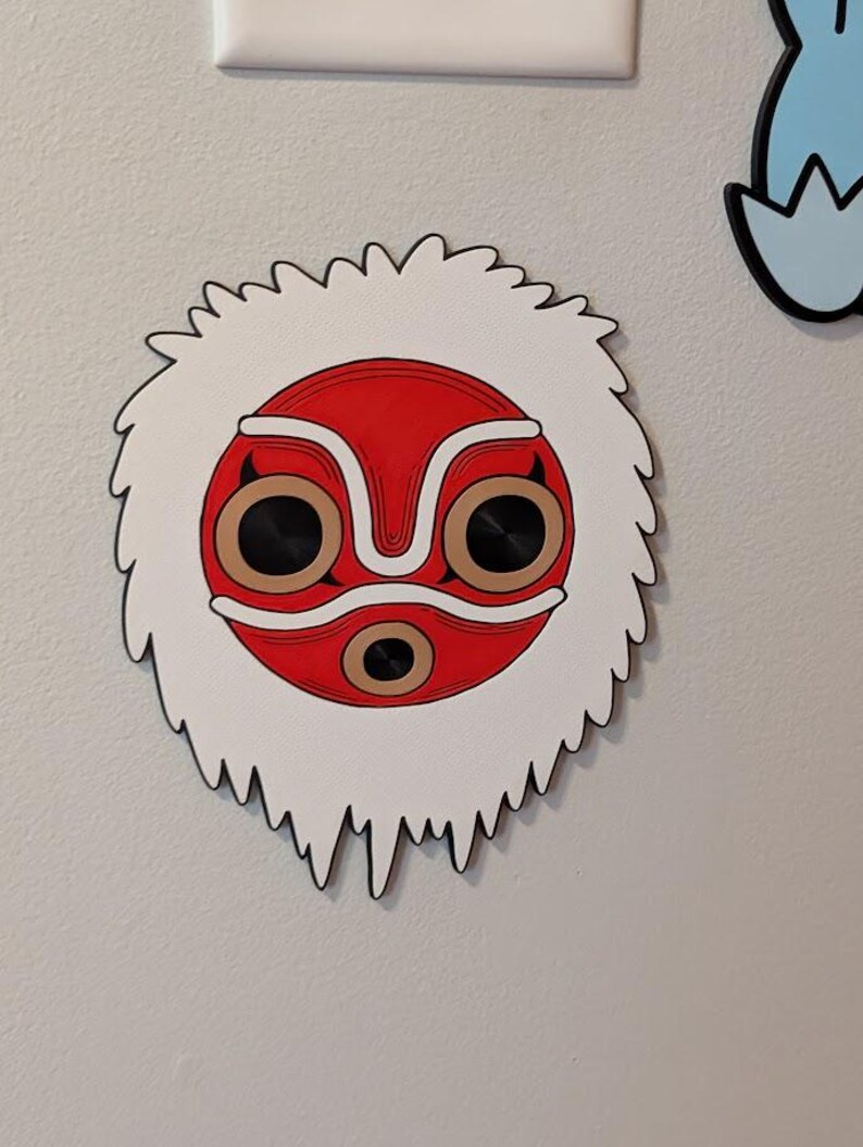 May include: A decorative wooden mask featuring a red face with white and brown accents, and black eye sockets. The mask is surrounded by a white, furry border with a jagged edge. The mask is mounted on a light gray wall.