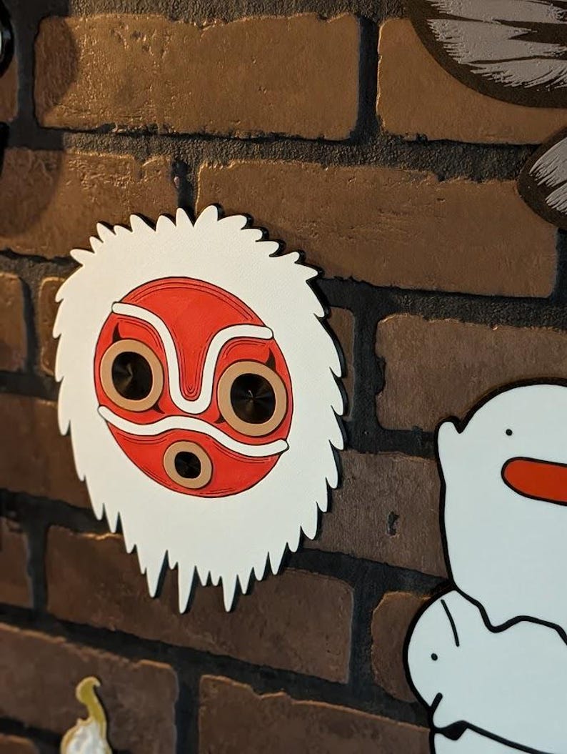 May include: A wooden wall decoration featuring a red and white mask with a jagged edge. The mask has a red face with brown and white details, and two dark circles for eyes. The mask is mounted on a brick wall.