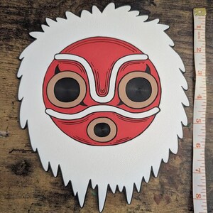 May include: A white and red mask with a stylized face design. The mask has a white, textured border and measures approximately 8 inches in diameter. The face features large, circular eyes and a symmetrical design.