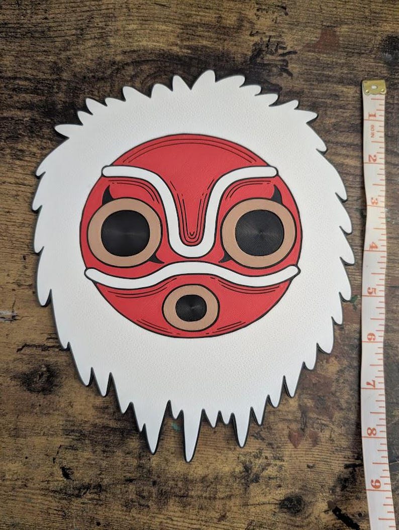 May include: A white and red mask with a stylized face design. The mask has a white, textured border and measures approximately 8 inches in diameter. The face features large, circular eyes and a symmetrical design.