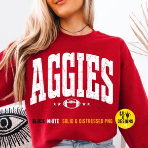 May include: A red sweatshirt featuring the word "AGGIES" in white, distressed lettering. A football graphic is below the word, flanked by stars. The text "BLACK WHITE SOLID & DISTRESSED PNG" is below the football graphic.