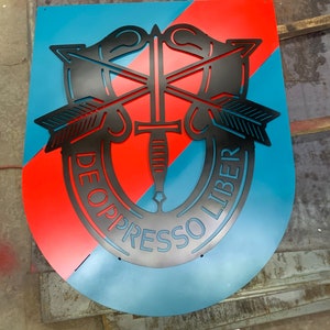 Peut inclure: Une découpe en métal noir de l'insigne des Forces spéciales de l'armée américaine, connu sous le nom de "béret vert". L'insigne représente un poignard ailé, des flèches et la phrase latine "De Oppresso Liber", qui se traduit par "Libérer les opprimés". L'insigne est monté sur un fond bleu et rouge.